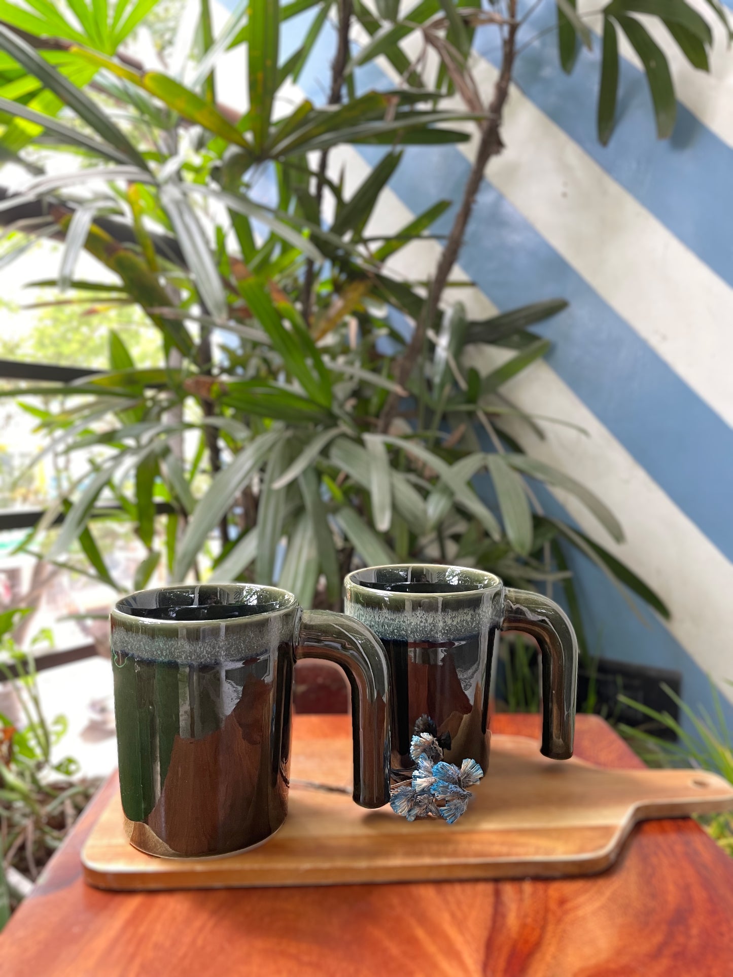 2 of tall green glaze ceramic mugs, shaded green at rim, ergonomically easy to hold half handles on wooden board with dried flowers in balcony. Shop traditional Indian ceramics