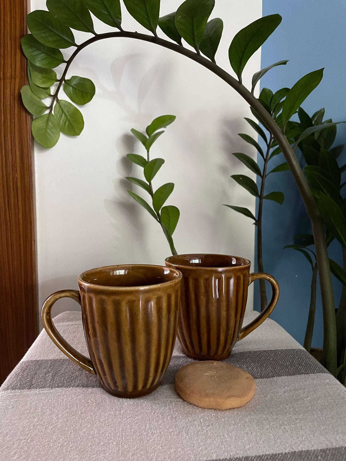 Pair of mustard glazed textured line pattern ceramic mugs on table with cookie, plants in backdrop. Shop traditional Indian ceramics.