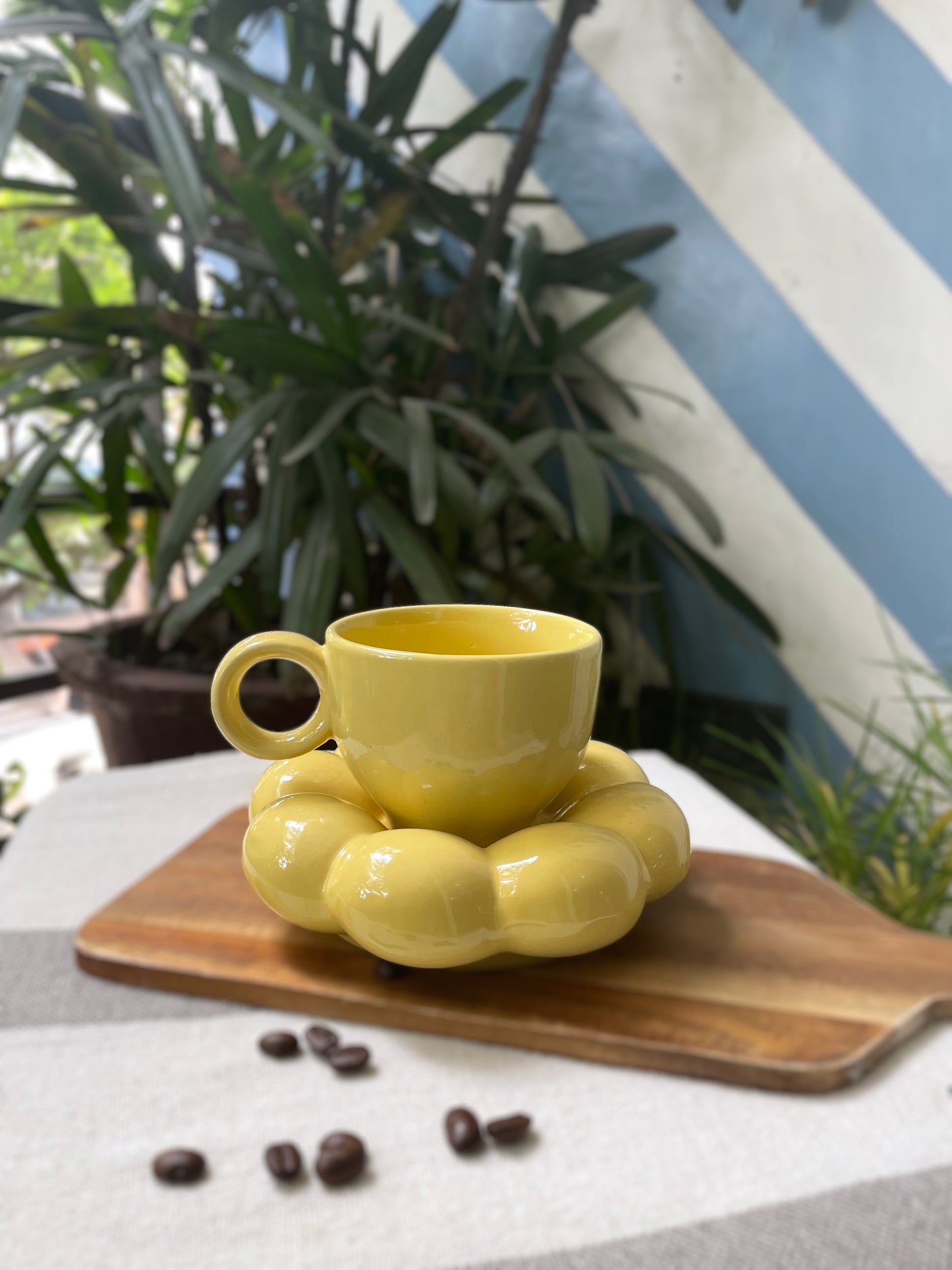 A set of yellow ceramic cup with round handle & flower-shaped saucer on wooden slab with coffee beans spilled around. Explore ceramics for Indian homes.