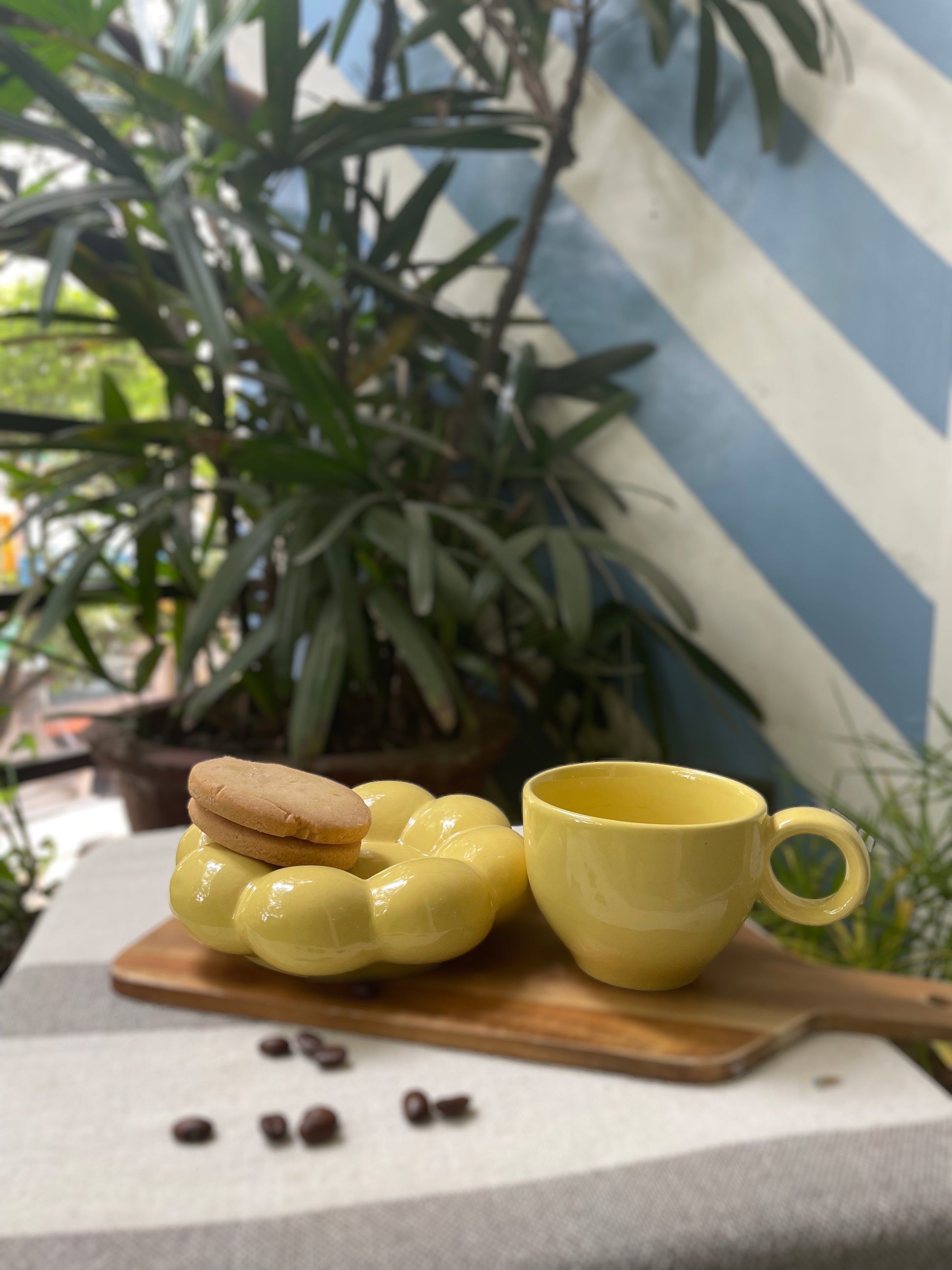 A yellow ceramic cup & flower-shaped saucer on wooden slab with spilled coffee beans and cookies, in outdoor setting. Shop handmade ceramics India.