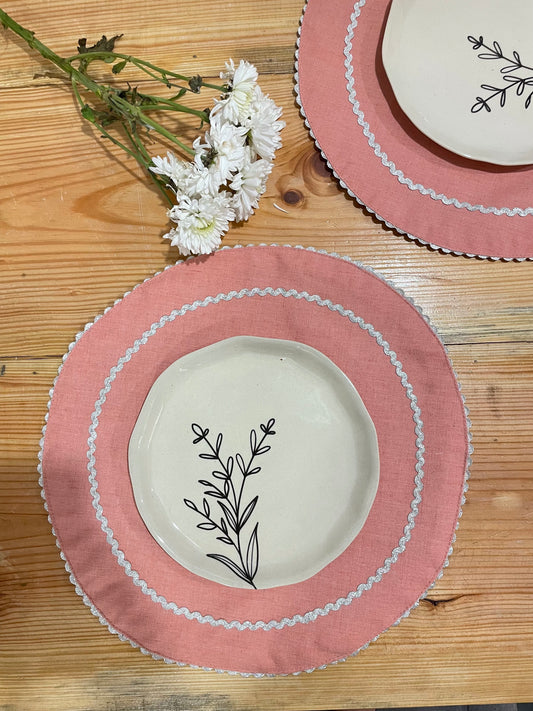 Pair of pink round table mats with silver scallop border and circle interior, round ceramic plates, and white flower. Buy tableware online in India