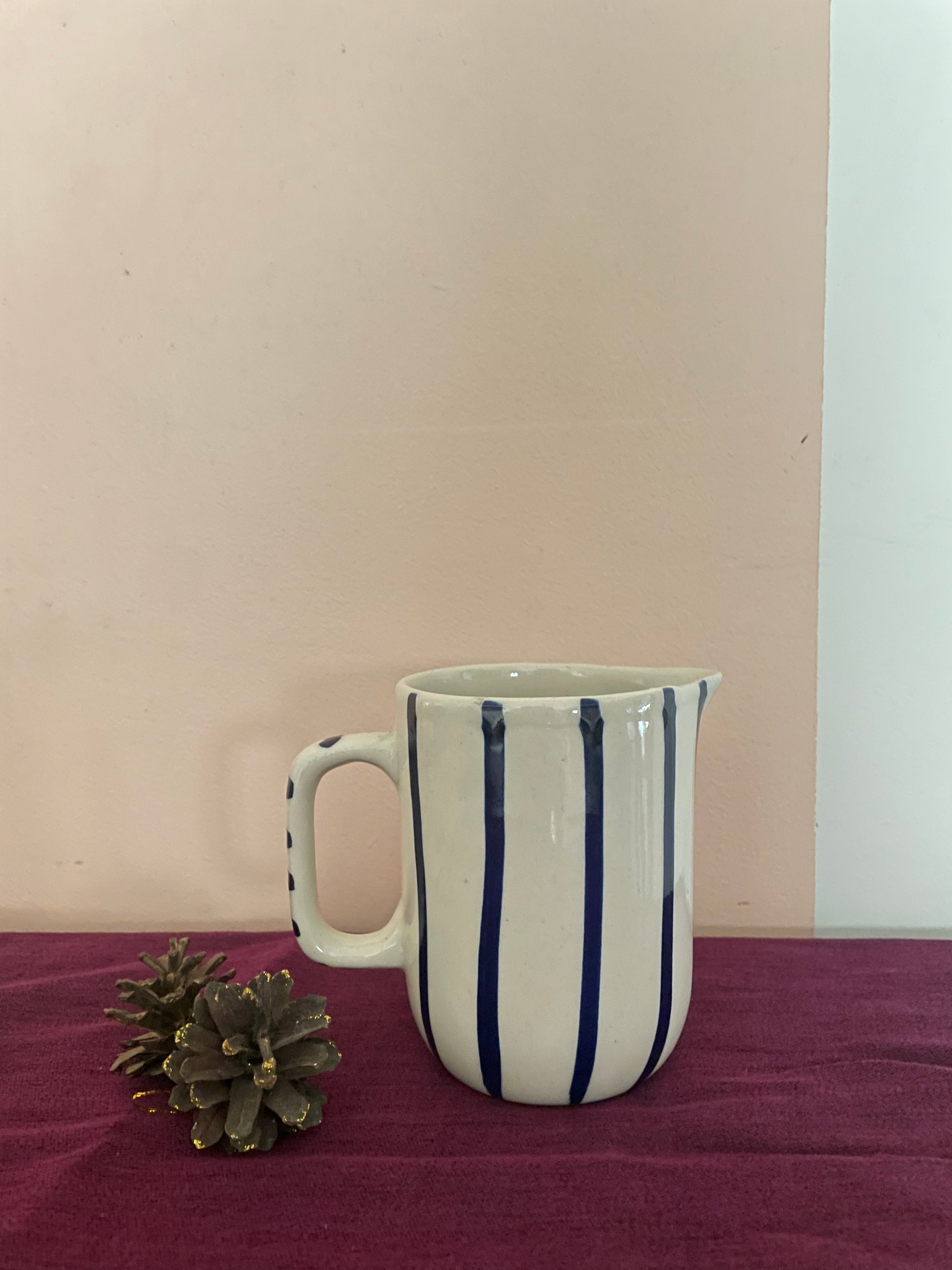 Small off-white ceramic jug with handle, hand-painted thin blue stripes kept on purple cloth with pine cones. Explore unique gifting ideas Bangalore.