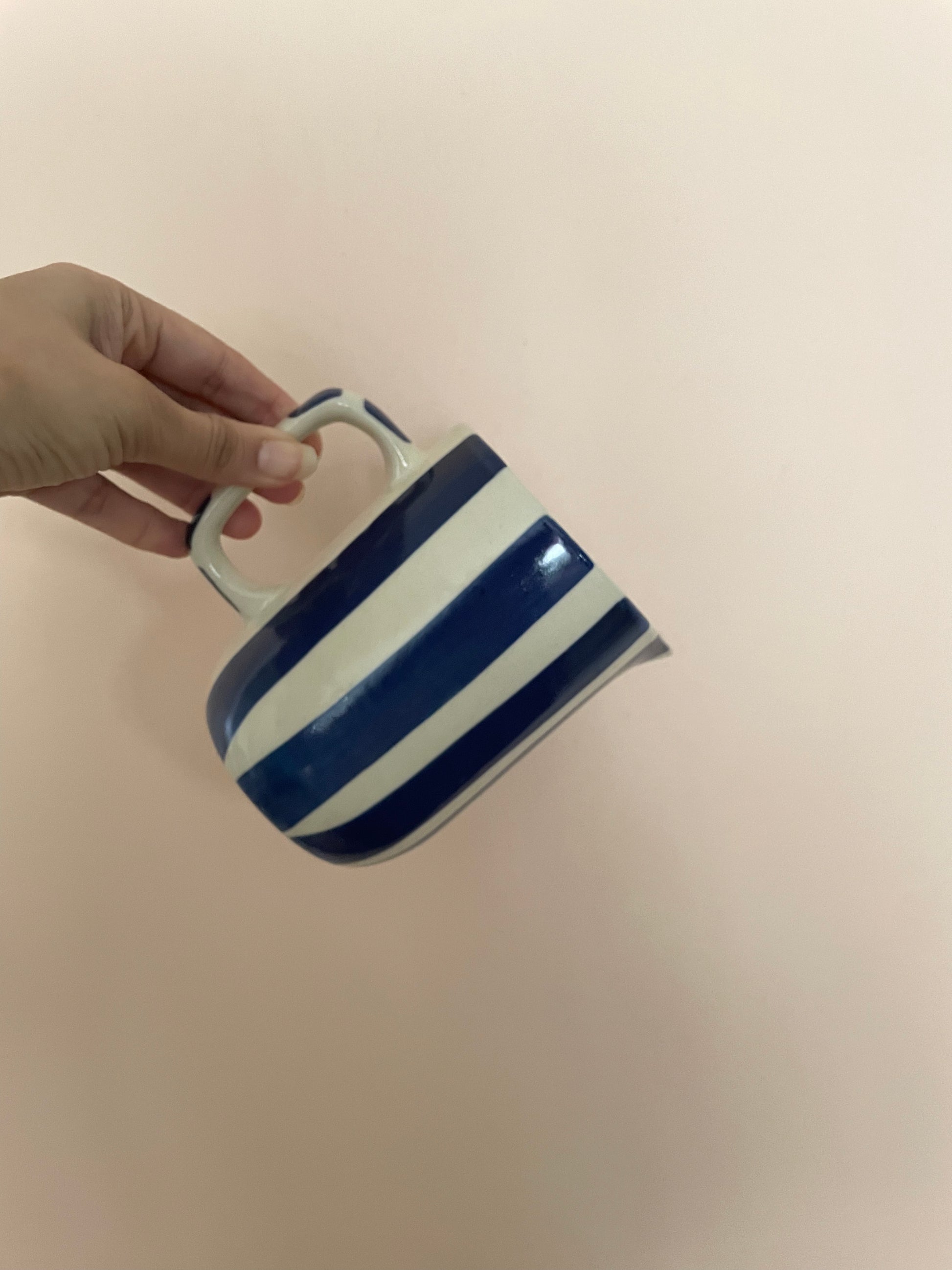 Hand holding small off-white ceramic jug with square-ish handle, blue hand-painted broad stripes against pink wall. Explore luxury ceramics Bangalore.