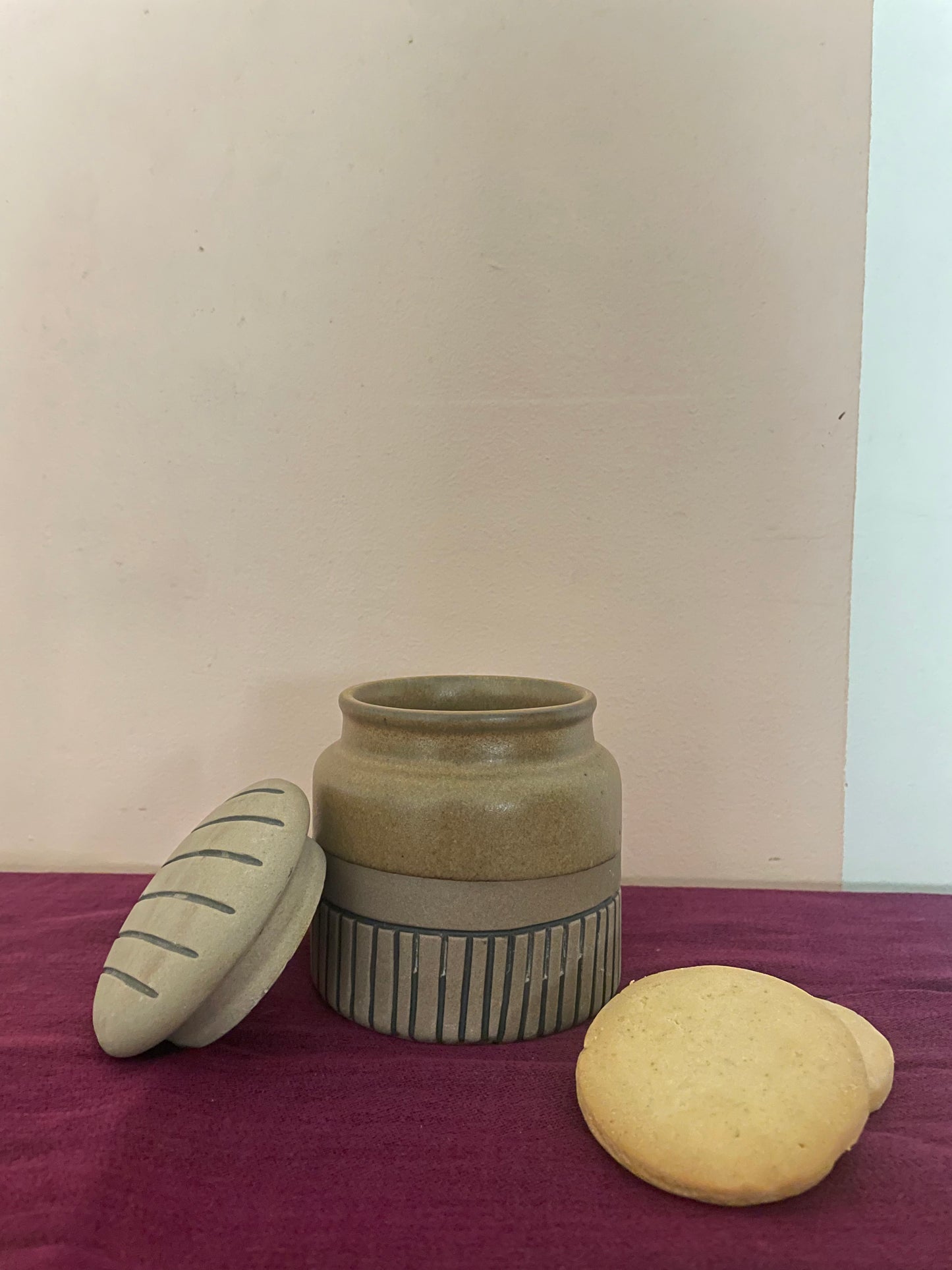 Medium mustard ceramic jar with brown bottom and black striped bottom, open round lid with stripes, beside cookies. Buy home decor gifts India.