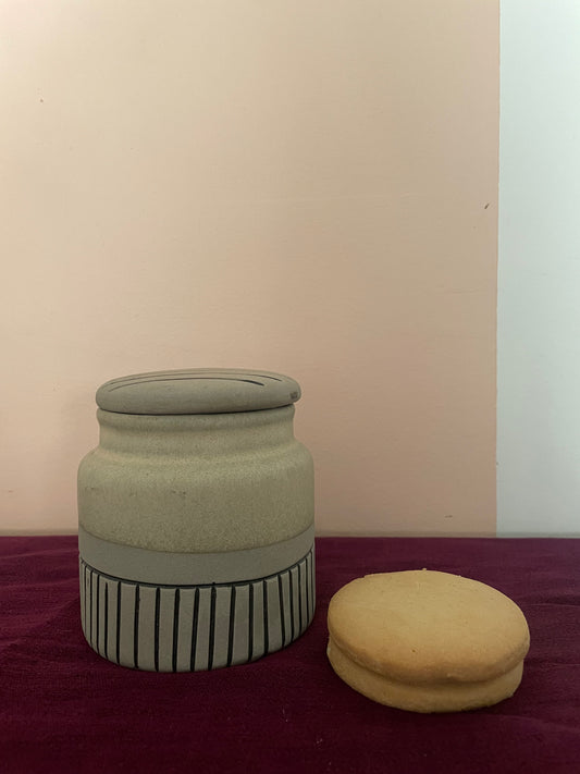 Medium stoneware ceramic jar in beige with grey and black stripes on bottom half, round lid with stripes, cookies beside it. Shop ceramics for Indian homes.