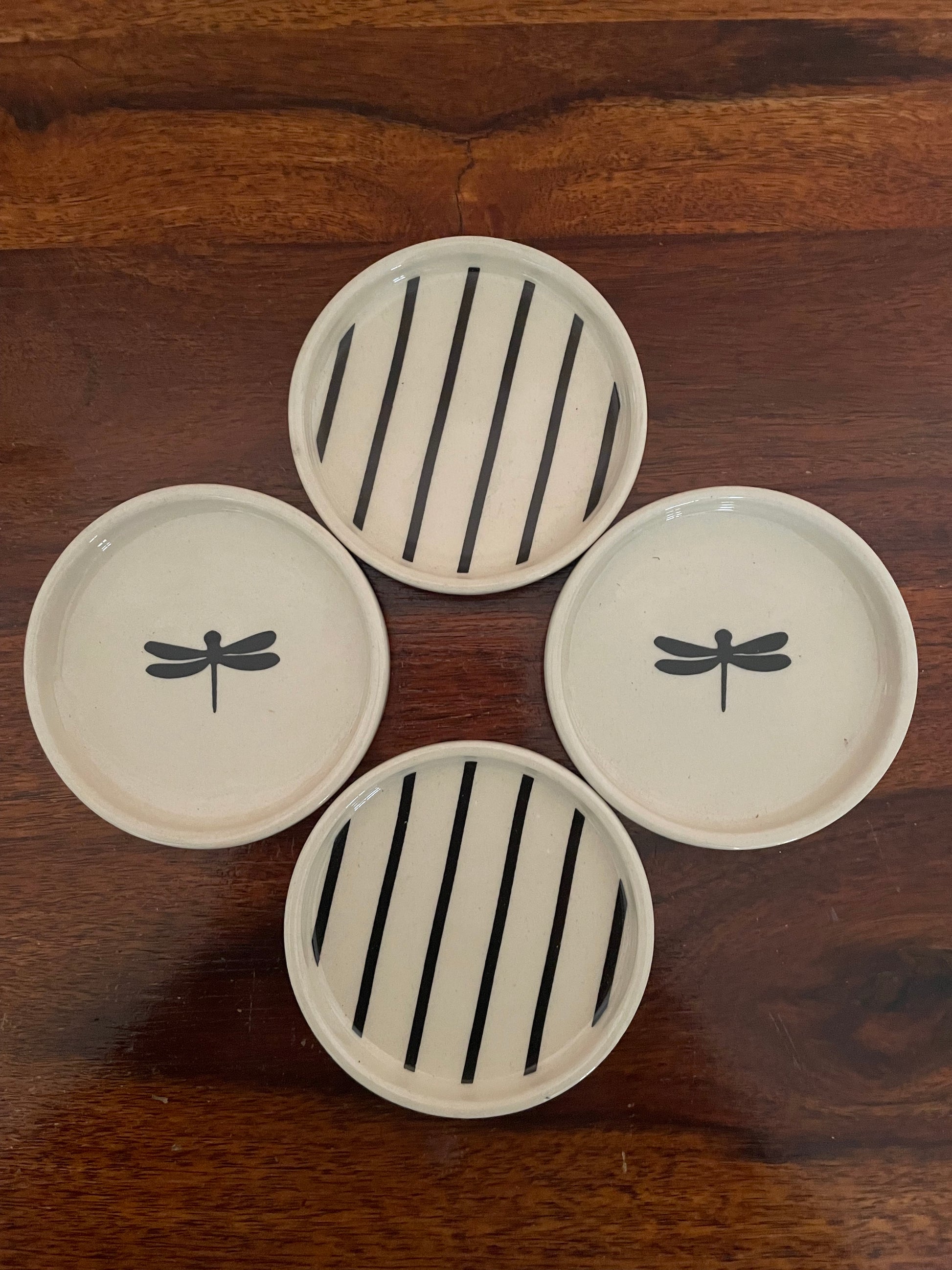 Top view: Set of 4 round off-white coaster, 2 with black dragonfly motifs and 2 with black stripes, on a wooden table. Explore unique gifting in India and Bangalore