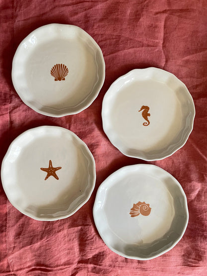 Set of 4 uneven hem small off-white ceramic plates with unique ocean-inspired designs on rusty cloth. Shop designer tableware India.