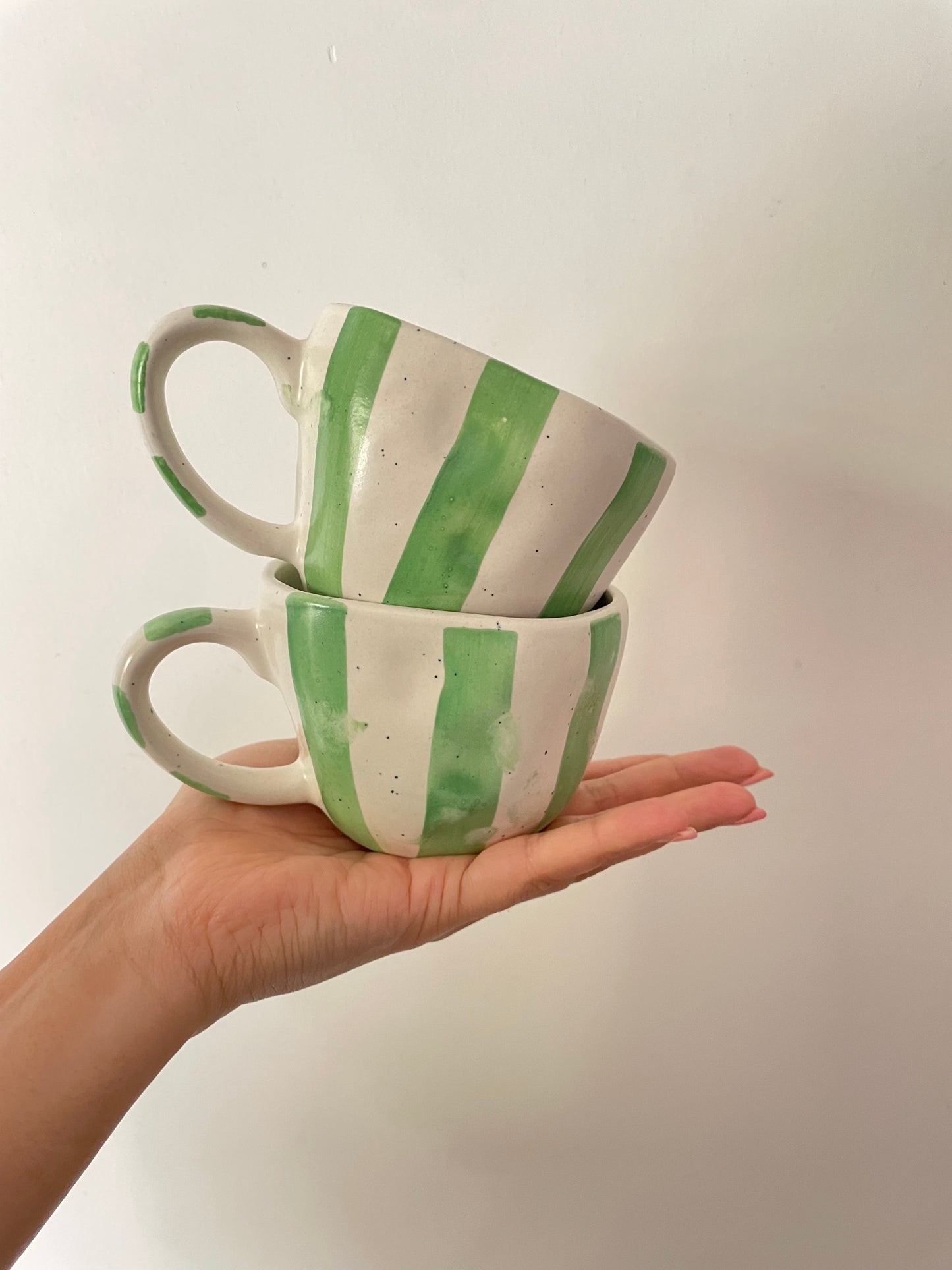 Pair of short uneven hem off-white ceramic mugs: green painted stripes on mug and handle, balancing on a lady's palm. Discover OYP's online ceramics store India.