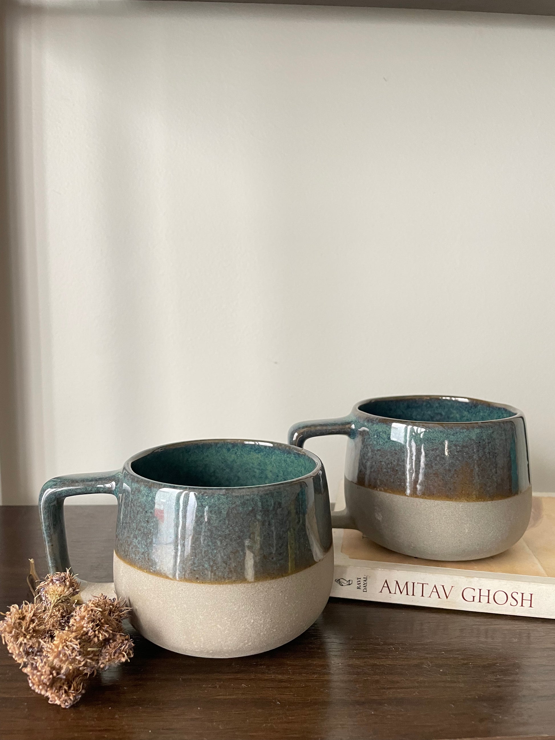 Pair of small ceramic mug: green glazed top, beige stoneware bottom, squareish handle. 1 on table with dried flowers, 1 on book. Shop ceramics for Indian homes.