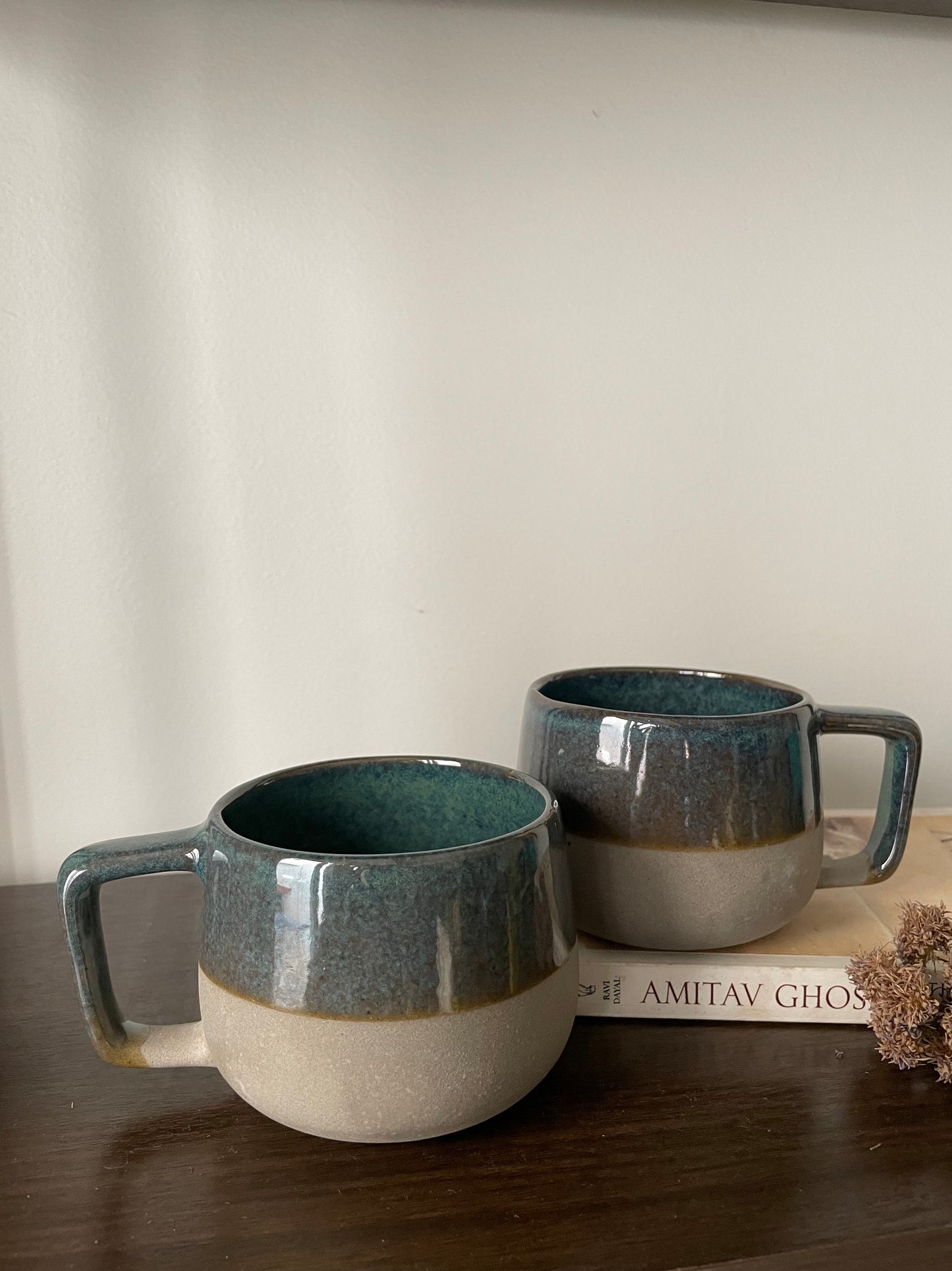 A pair of ceramic mug, green top glaze, stoneware bottom half. One on wooden table, one on book with dried flowers. Explore best ceramics stores in Bangalore