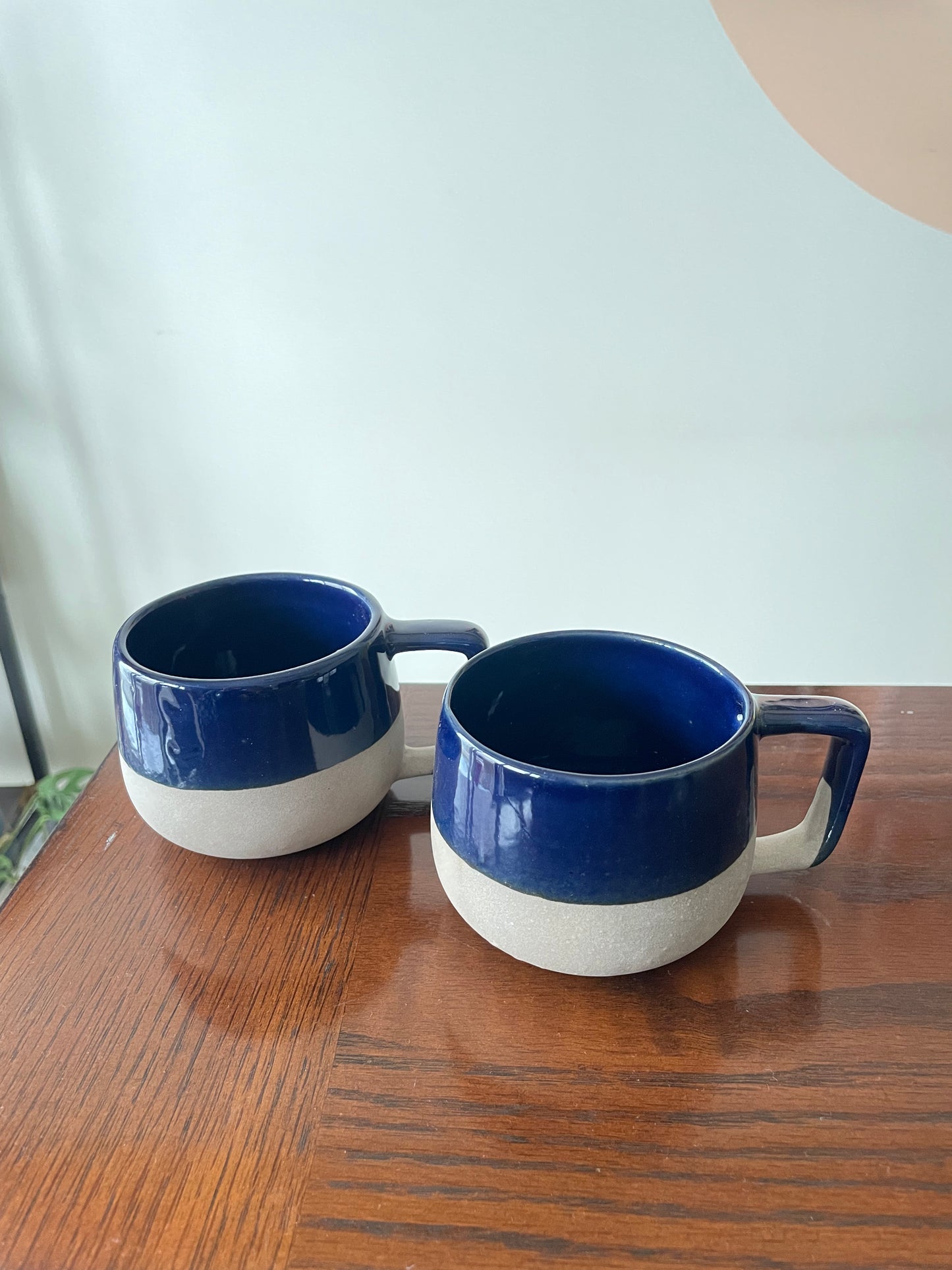 Pair of short, broad ceramic mugs, unique squareish handle, blue glaze on top half, stoneware finish on bottom half, on wooden table. Explore best ceramics stores in Bangalore