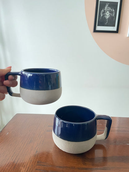 Hand holding up short, broad ceramic mug, other on table. Top half glazed blue, half beige, including handles. Shop unique mugs online Bangalore