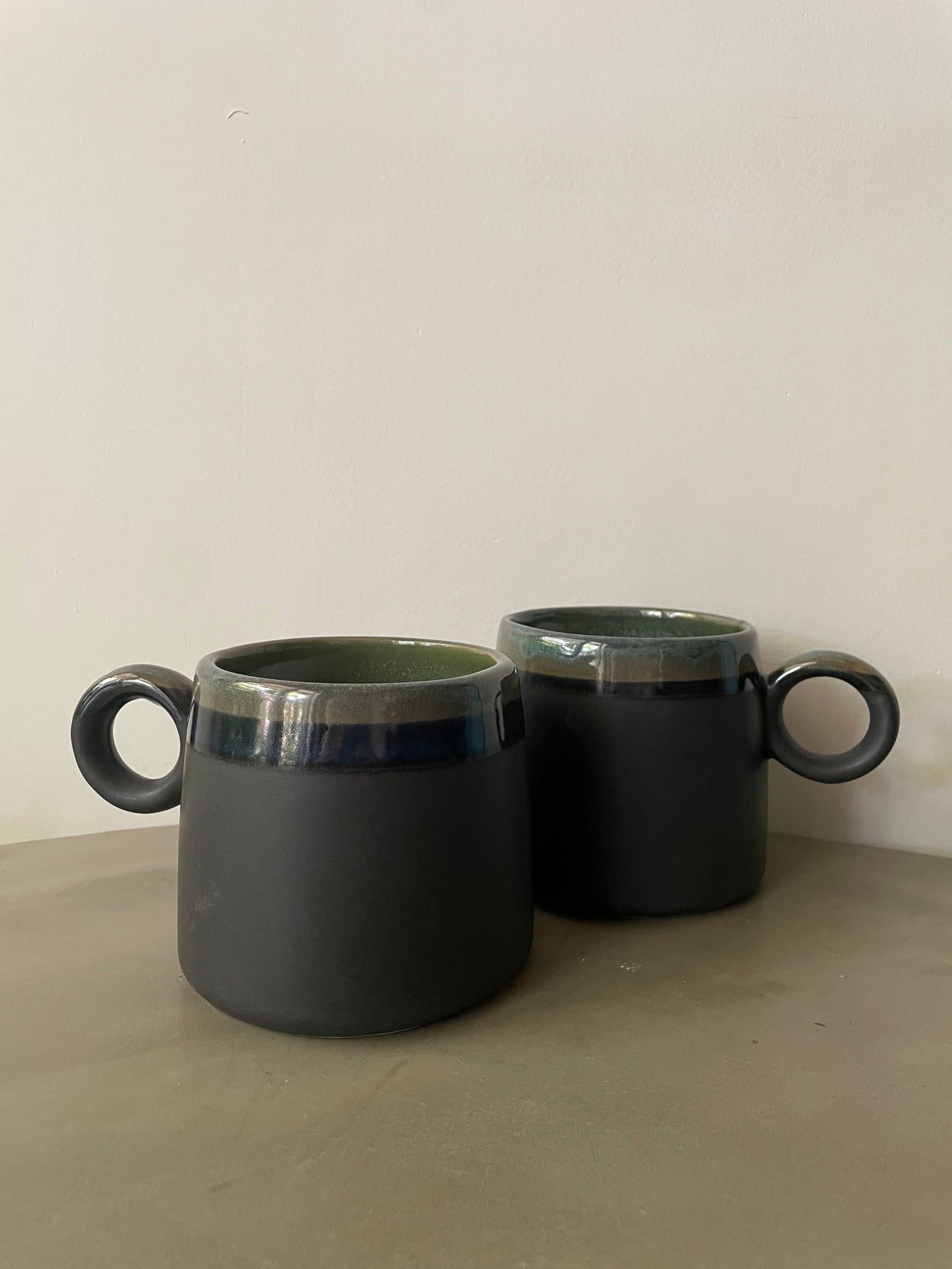 Pair of medium rounded ceramic mugs with round handles, dual glaze finish in green tones and black, on a cement coffee table. Shop artistic ceramics in Bangalore