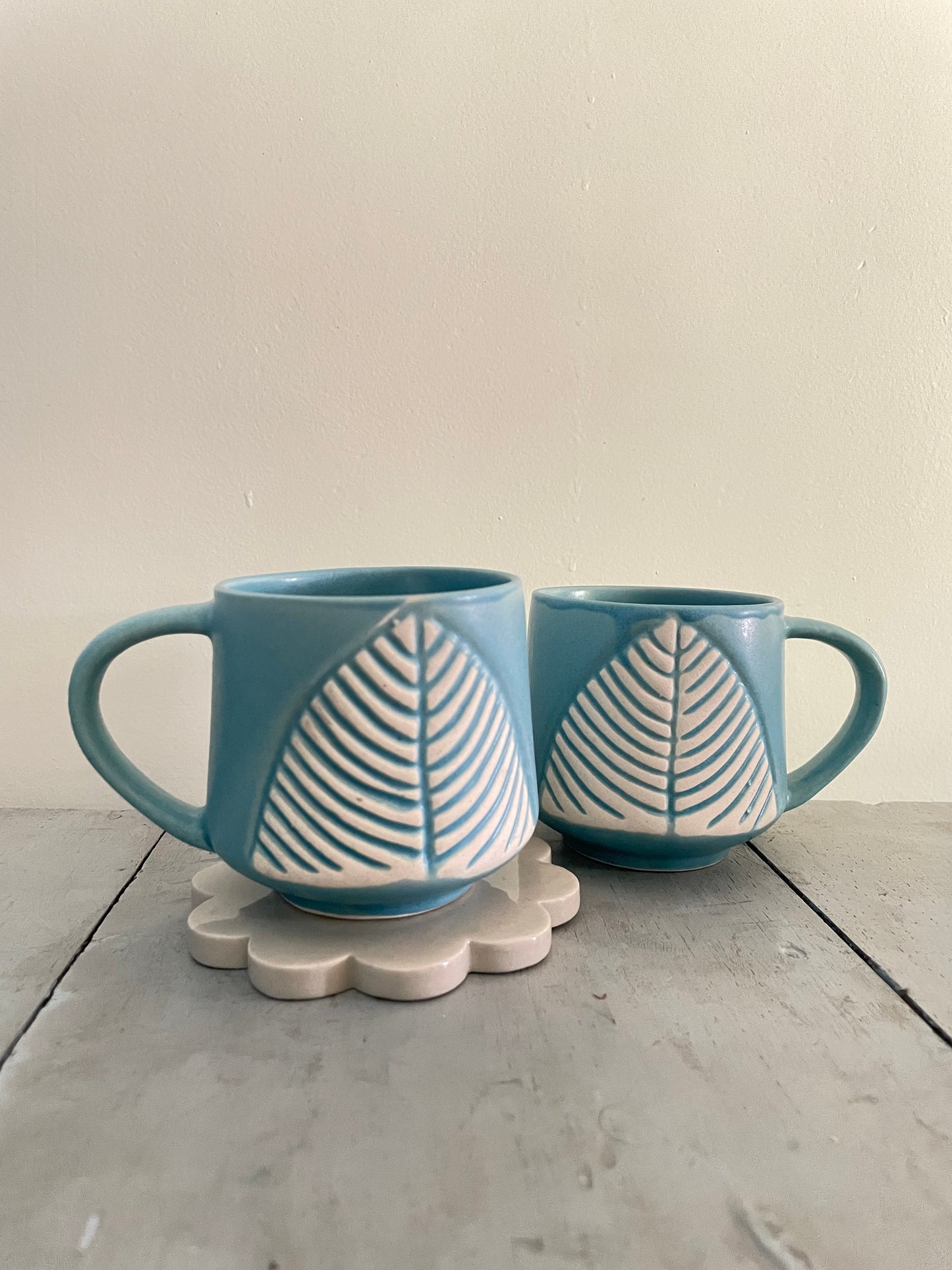 Pair of mini teal ceramic mug with big handle and off-white leaf motifs, one on a white ceramic flower coaster, other diagonally behind. Shop handcrafted ceramics in India