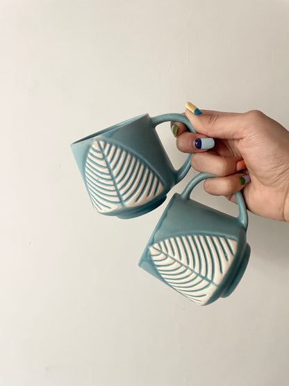 Hand with vibrant painted nails holding a pair of mini teal mugs with broad handles and large white leaf motif. Explore unique mugs in Bangalore.