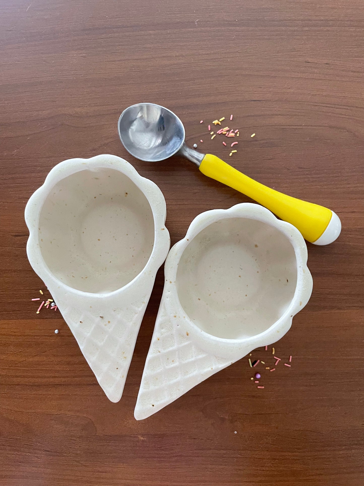 2 white ceramic waffle cone-shaped ice cream bowls with scallop rims and brown freckles on a wooden table, sprinkles, and scooper.