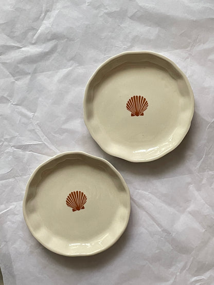 Set of 2 small uneven hem off-white ceramic plates with orange sea shell motif on white sheet. Shop ceramics for Indian homes.