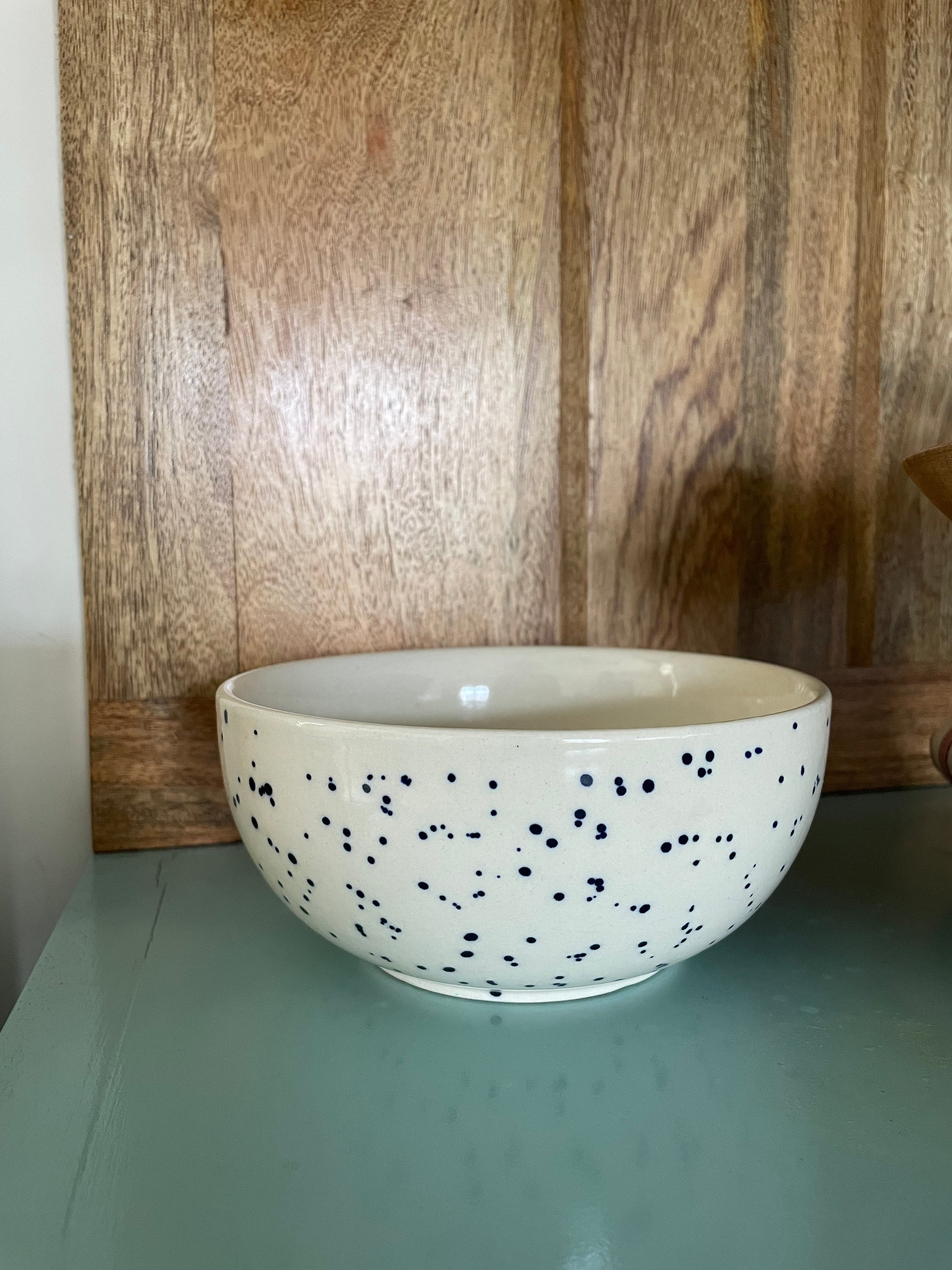 Medium ceramic bowl white glaze with blue splatter spots on teal blue table, wooden backdrop. Shop handcrafted ceramics in India at Oh Yay Project