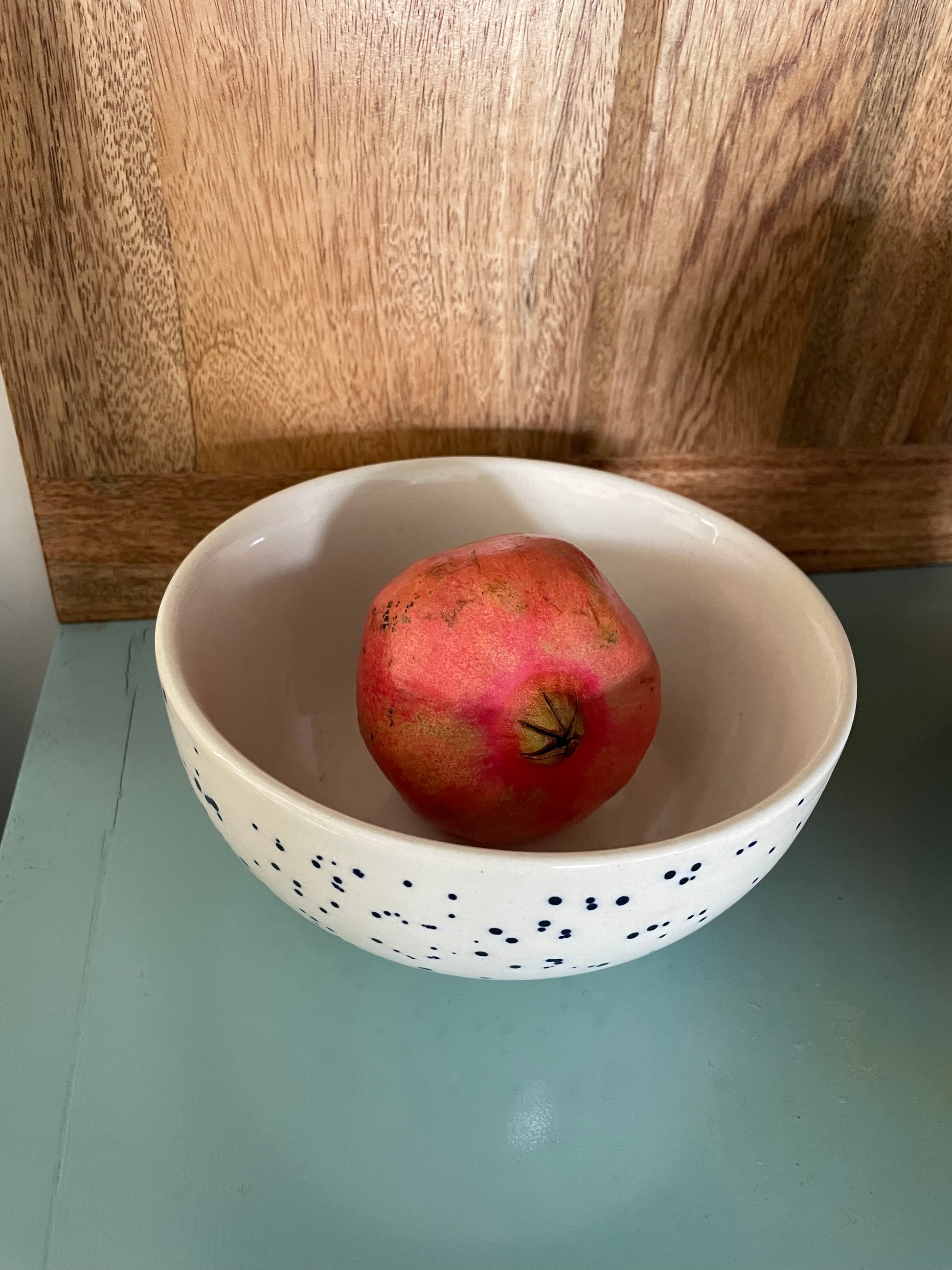 White glazed ceramic bowl on teal table, wooden backdrop, blue splatter spots. Inside white glaze, with pomegranate. Buy online for your tableware in India.