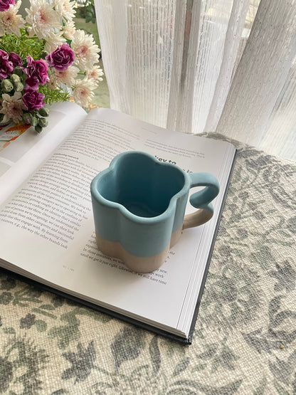 A ceramic mug with 4-petal shape rim, double-curve handle, blue and grey stoneware finish, placed on a book with flowers. Buy artistic ceramics in India.