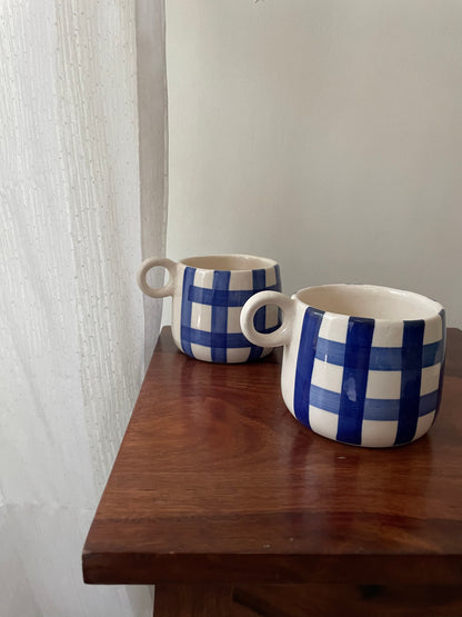 A pair of cute short small rounded ceramic mugs with blue hand-painted checks, small round handle, placed on table. Buy handcrafted ceramics in India.