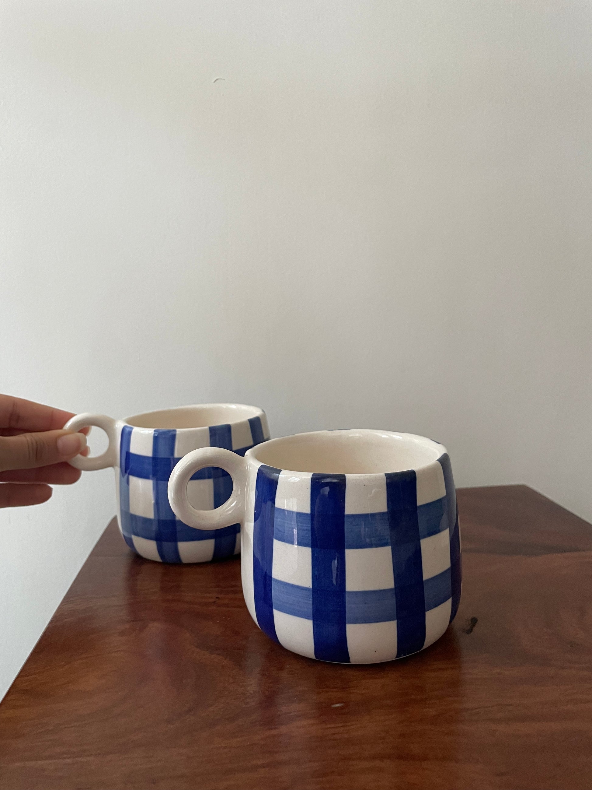 A pair of short cute ceramic mugs with hand-painted blue checks, small round handle, delicately held, on table. Buy handmade mugs in India.