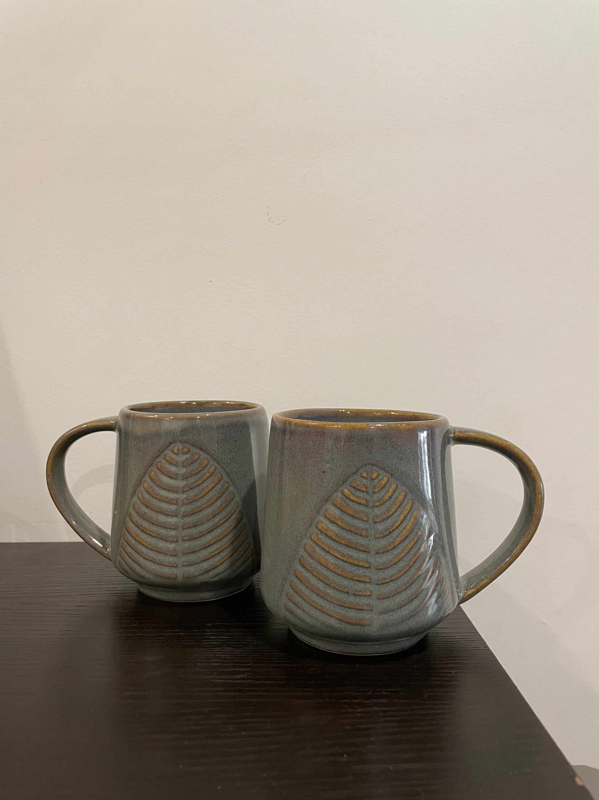 A pair of greyish-green big mugs with tonal leaf motif, wide handle for easy use, on table. Buy affordable ceramics in India.