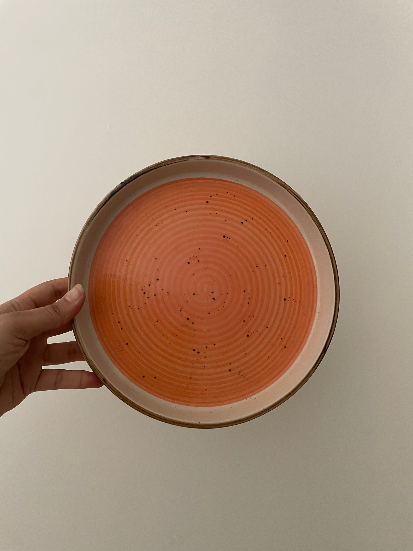 Hand holding upright a ceramic orange plate with white border, brown freckle design, and brown rim. Buy tableware online in India.