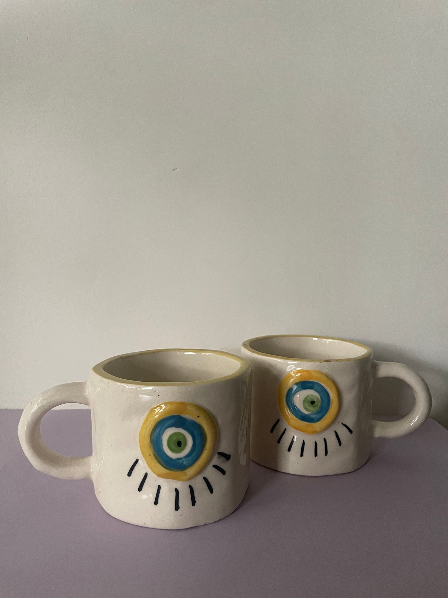 2 medium off white ceramic mugs with 3d embossed evil eye in yellow, blue & green.Shop designer tableware Bangalore