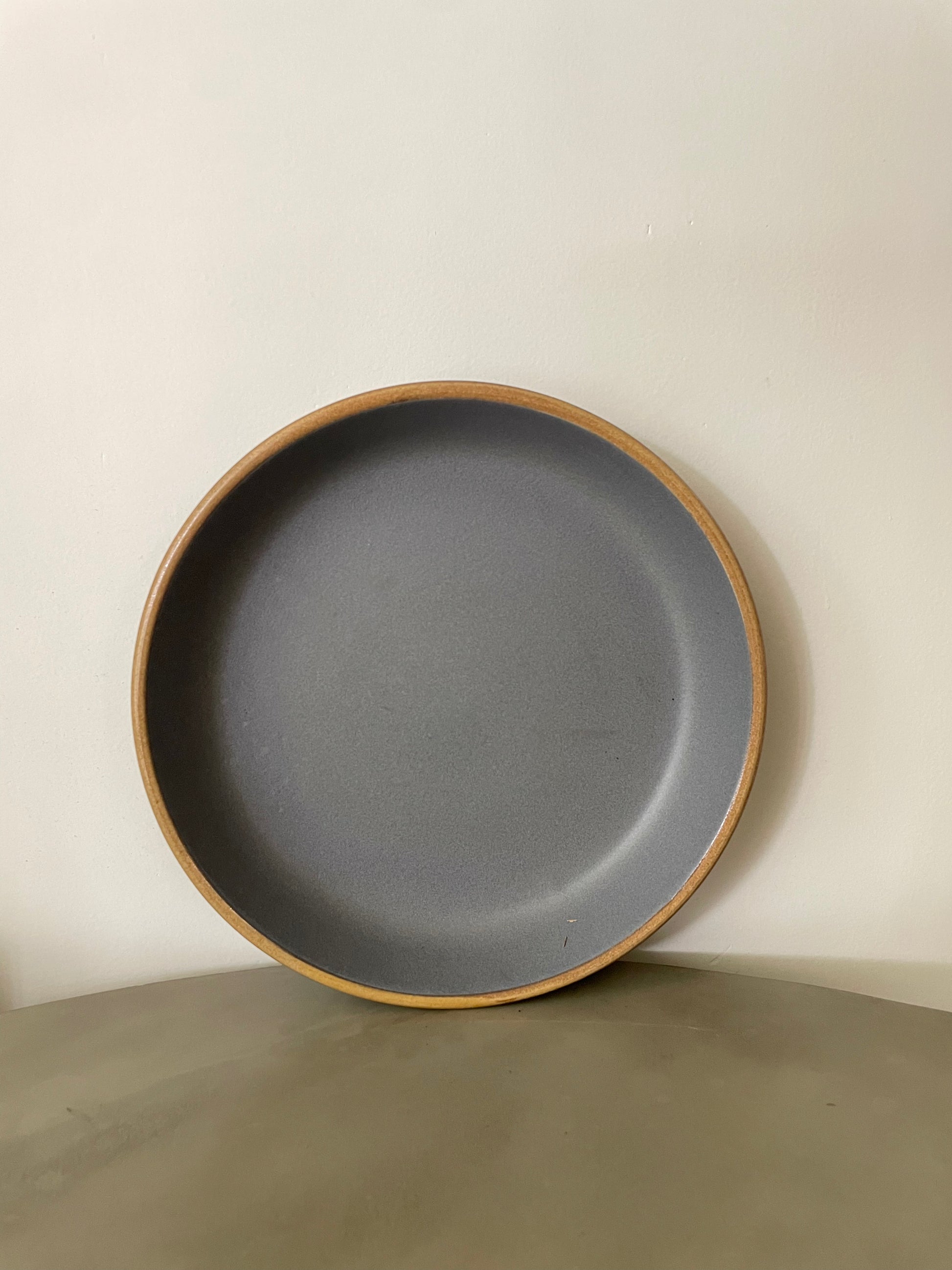 A large round ceramic plate with grey stoneware finish and mustard rim, placed upright against white wall on a cement table. Buy traditional Indian ceramics.