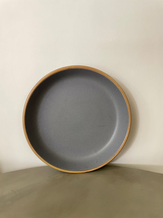 A large round ceramic plate with grey stoneware finish and mustard rim, placed upright against white wall on a cement table. Buy traditional Indian ceramics.