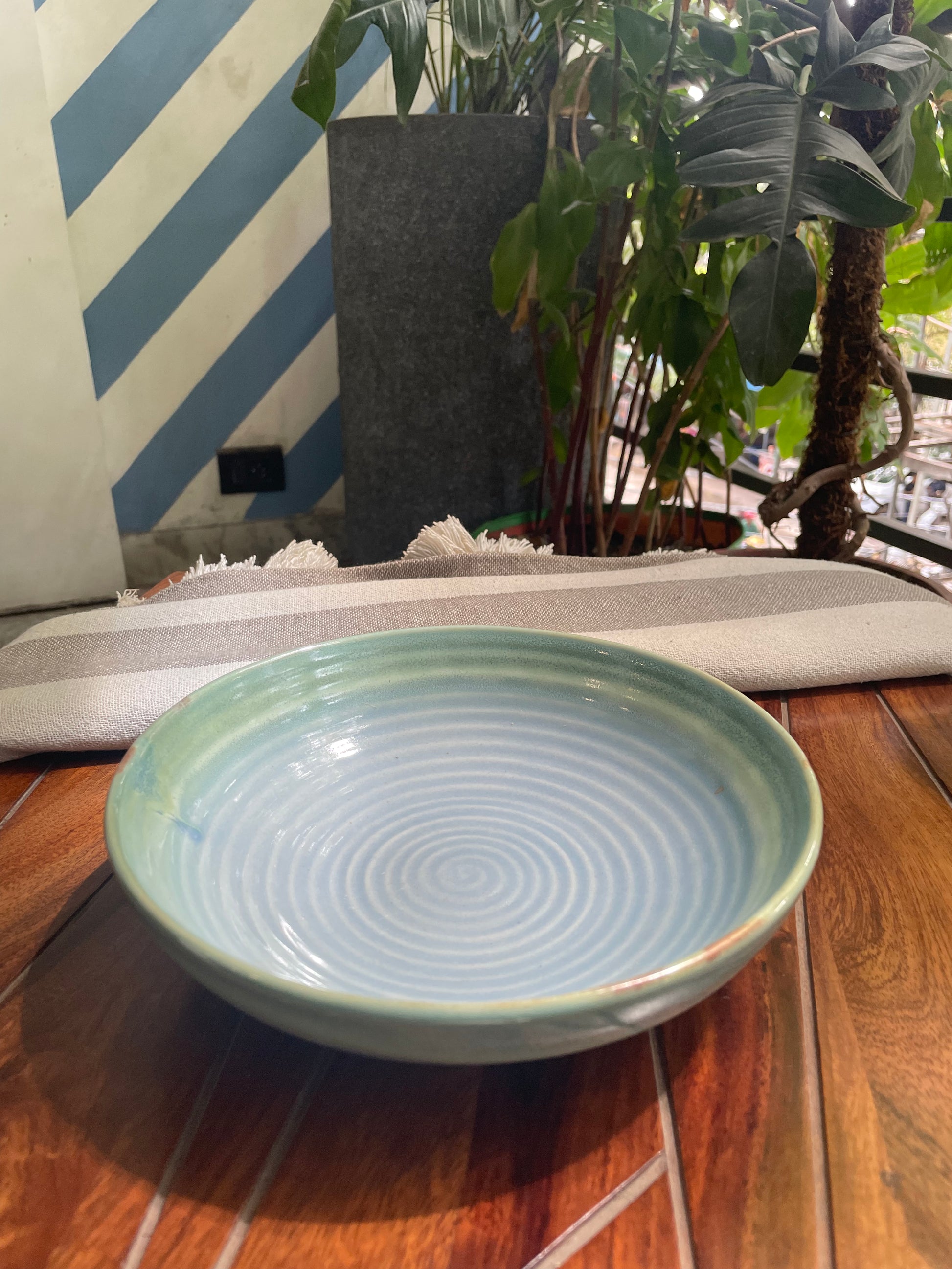 Angular view of ceramic shallow bowl in shades of blue glaze with hint of green, circular pattern inside, in outdoor setting. Discover handcrafted ceramics in Bangalore.