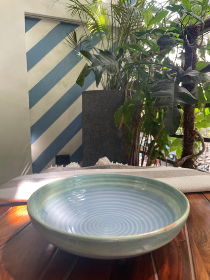 A bluish-green glazed shallow ceramic bowl with circular pattern inside on a wooden table in an outdoor setting. Shop custom ceramics in Bangalore.