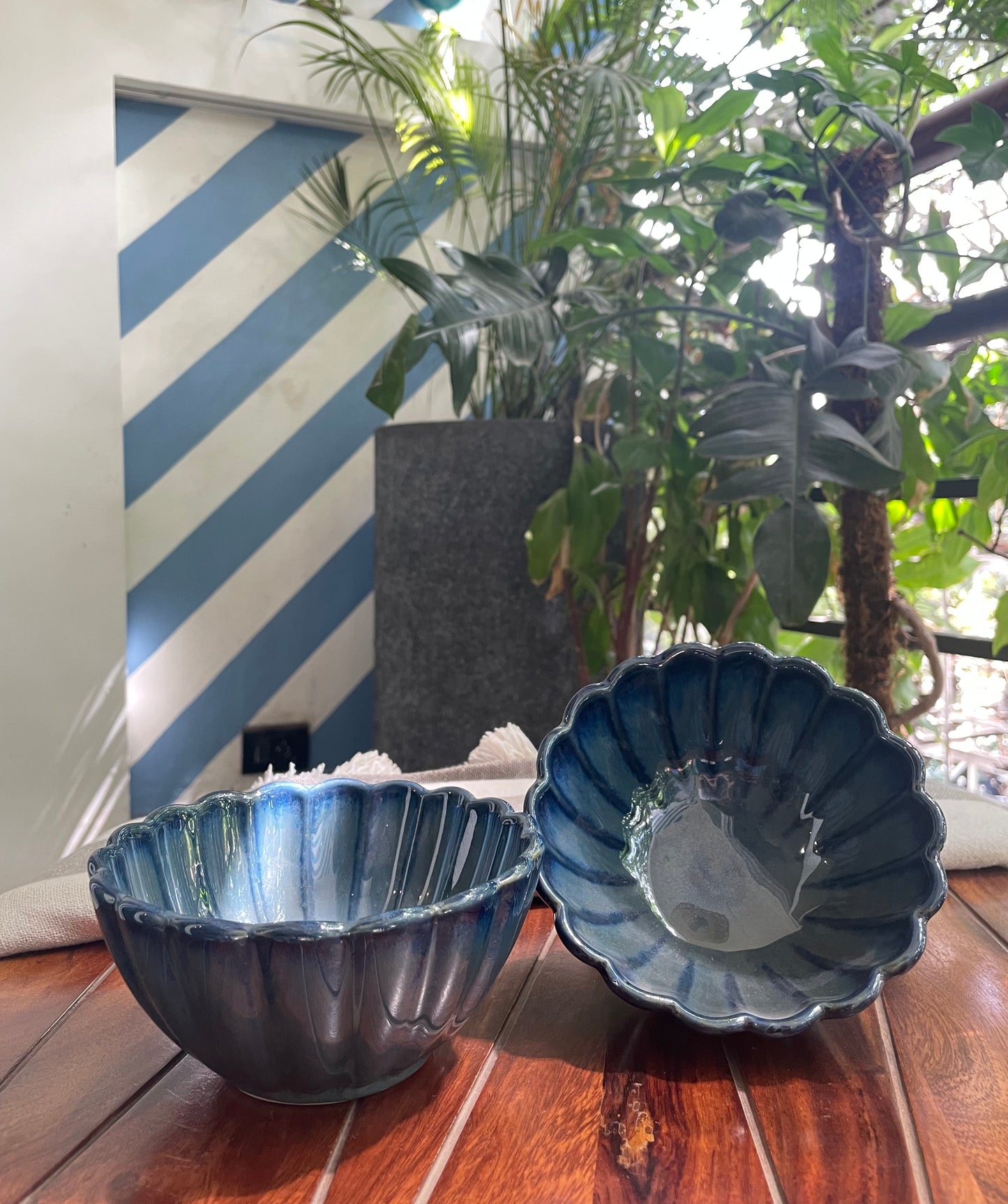 Pair of flower-shaped deep ceramic bowls in blue glaze, one on table, the other tilted, placed in outdoor setting. Buy affordable ceramics in India.