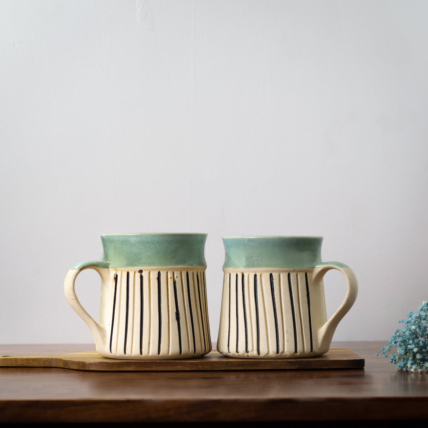 Pair of ceramic mugs with handle: light blue top rim, white stoneware finish with black stripes, on wooden slab with blue dried flowers. Buy designer tableware Bangalore.