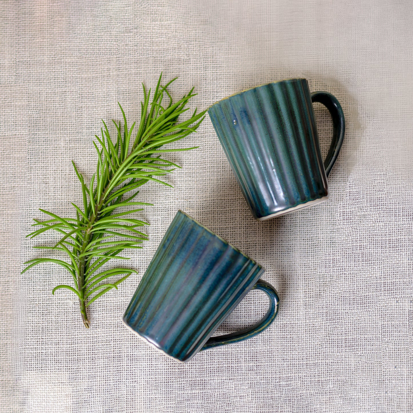 Top shot of 2 ceramic mugs with green glaze, ribbed detailing, handle, green leaf next to them on jute background. Buy kitchenware India.