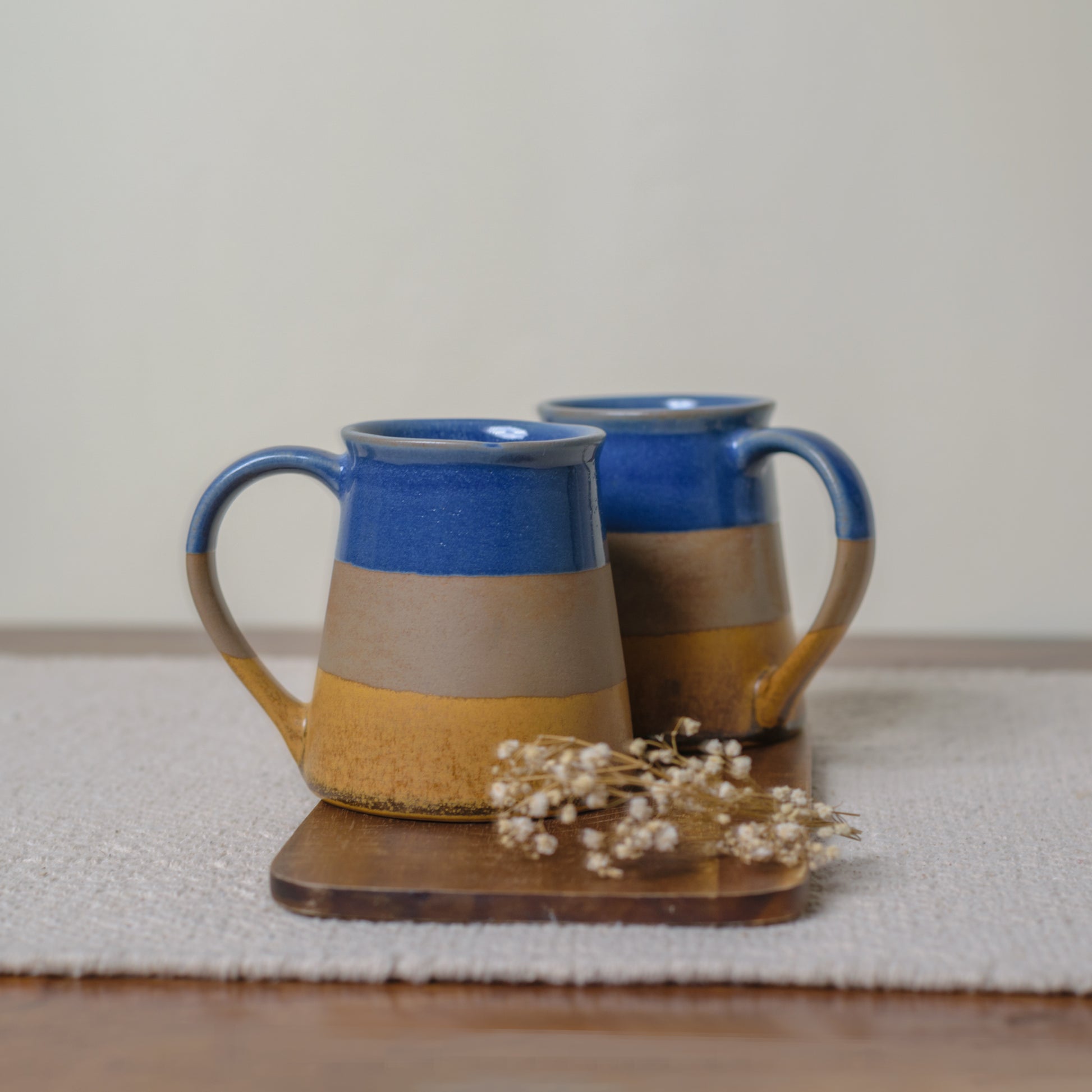 Pair of tall ceramic mug with handle, broad to narrow mouth, 3 glaze finish (blue, brown & mustard), on wooden slab, white dried flowers. Buy traditional Indian ceramics.