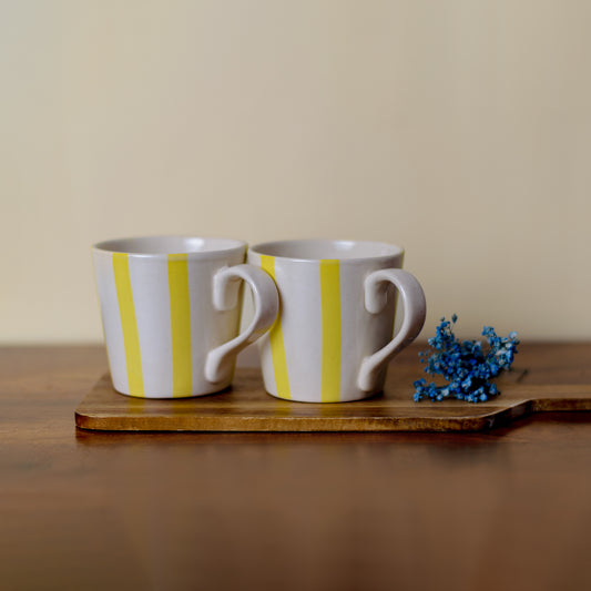 A pair of mid-sized off-white ceramic mugs with handles, hand-painted yellow stripes, on wooden slab with blue dried flowers.