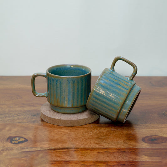 Pair of green glaze ceramic cups with squarish handles, subtle line detailing. One on MDF round coaster, other tilted. Shop home decor gifts India.