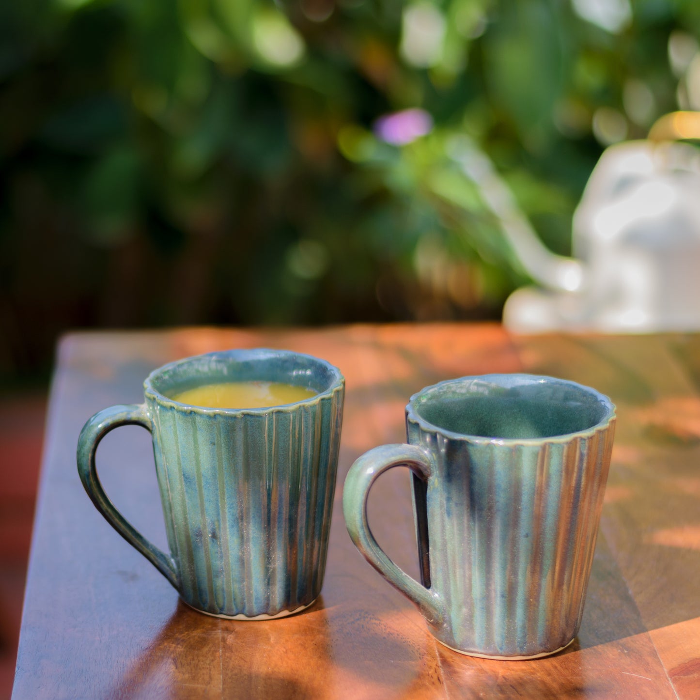 Set of green glaze ceramic mugs with tonal ribbed design and handle, one with juice, on wooden table with blurry plants. Shop artistic ceramics Bangalore.