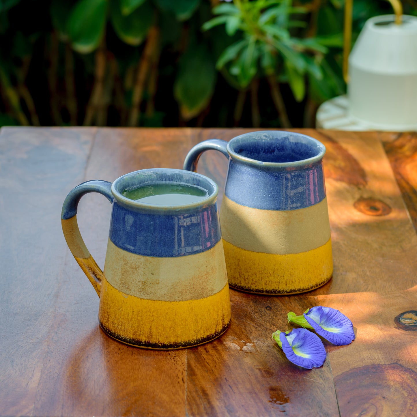 Pair of ceramic mugs with narrow rim, glazed in blue, brown & mustard stripes, one with juice, outdoor setting on wooden table with violet petals. Buy kitchenware India.