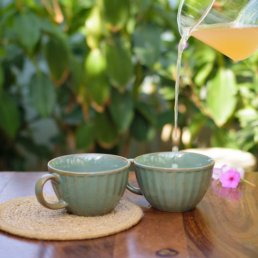 Pair of broad round green ceramic cups with handle, tonal ribbed detailing, 1 over jute mat, 1 with orange juice, blurry plant background. Explore unique gifting ideas Bangalore.