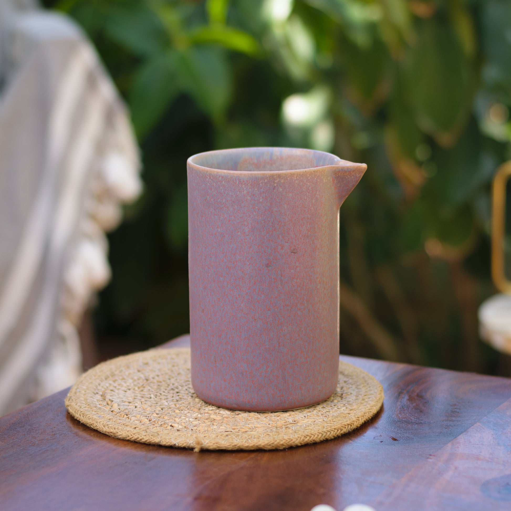 A large rounded ceramic jug with spout in mauveish purple glaze with blue hints on round jute mat, wooden table, plant in background. Shop ceramics for Indian homes.