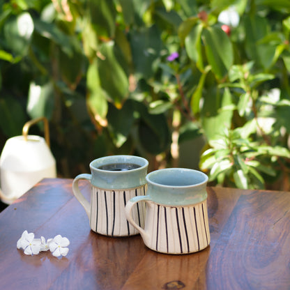 Set of 2 ceramic mug, blue glaze top rim, white stoneware finish with black stripes. One with coffee, one empty, on wooden table with white flowers. Shop handcrafted ceramics India.