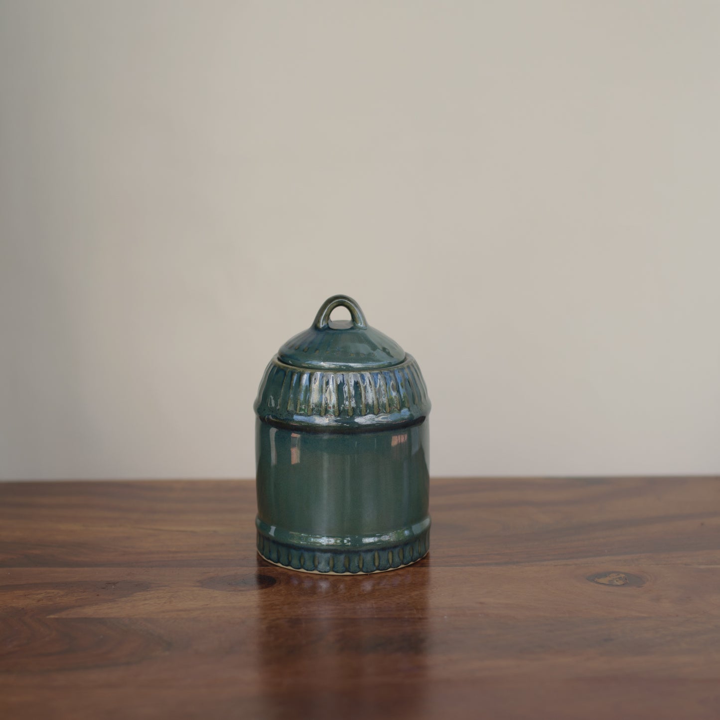 Medium ceramic jar in green glaze with dome-like lid, loop handle, tonal self-design placed on wooden table. Shop traditional Indian ceramics.