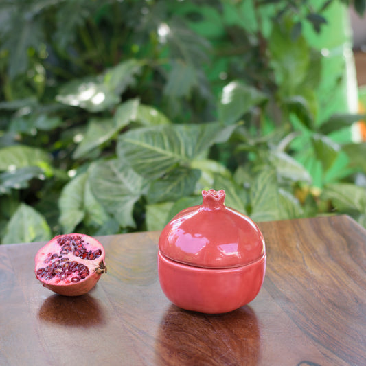 A medium red pomegranate-shaped jar with lid on wooden table, half-cut pomegranate, plants in background. Shop ceramics for Indian homes.