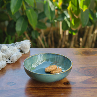 A green glazed ceramic bowl with 2 biscuits on a wooden table in an outdoor setting. Explore the best ceramic stores in Bangalore.