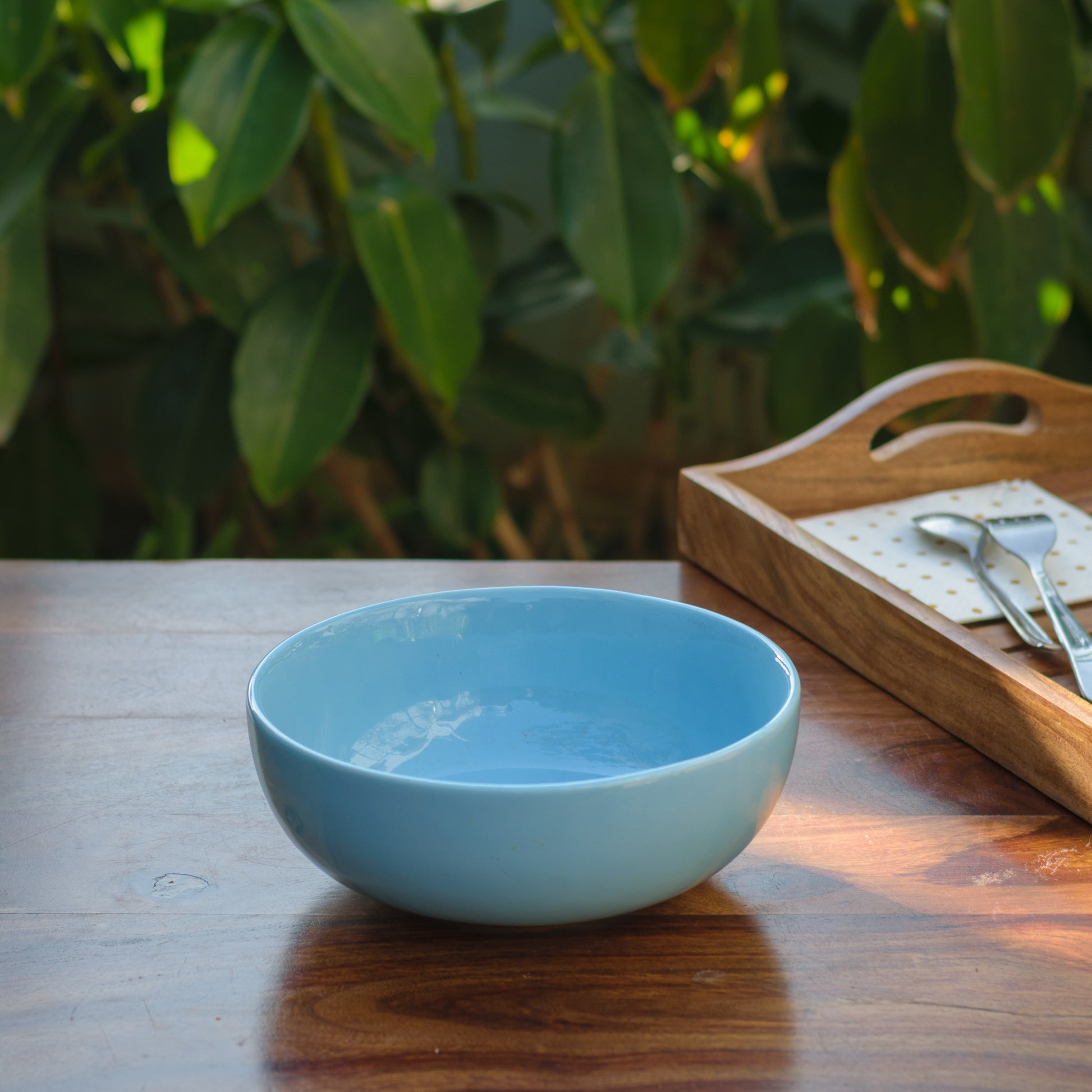 A solid sky blue glazed serving bowl on a wooden table with a tray of cutlery and napkin. Explore ceramics for Indian homes.