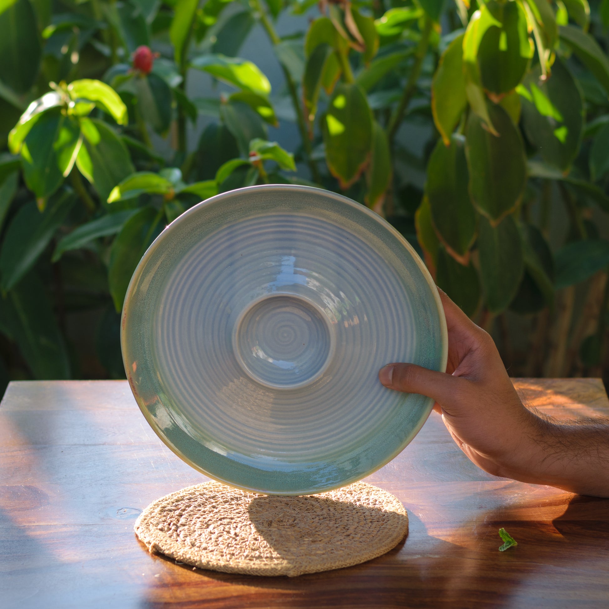 A hand holding a large round light blue ceramic platter with dip in middle, on a jute mat over a wooden table with plants. Buy ceramics for Indian homes.