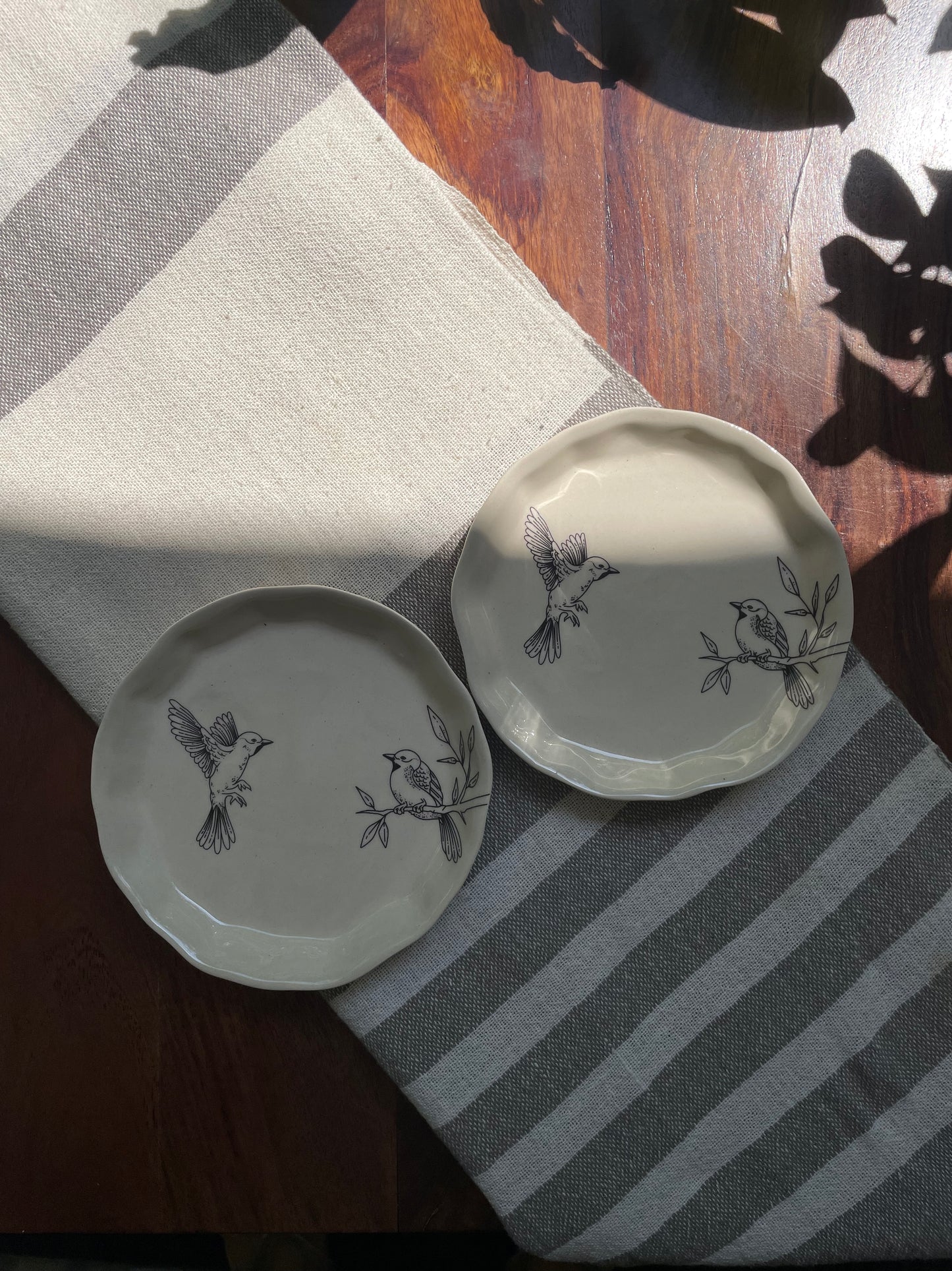 Top view of 2 small off-white ceramic plates with bird designs on a striped runner, with a wooden table and leaf shadow. Buy ceramics for Indian homes
